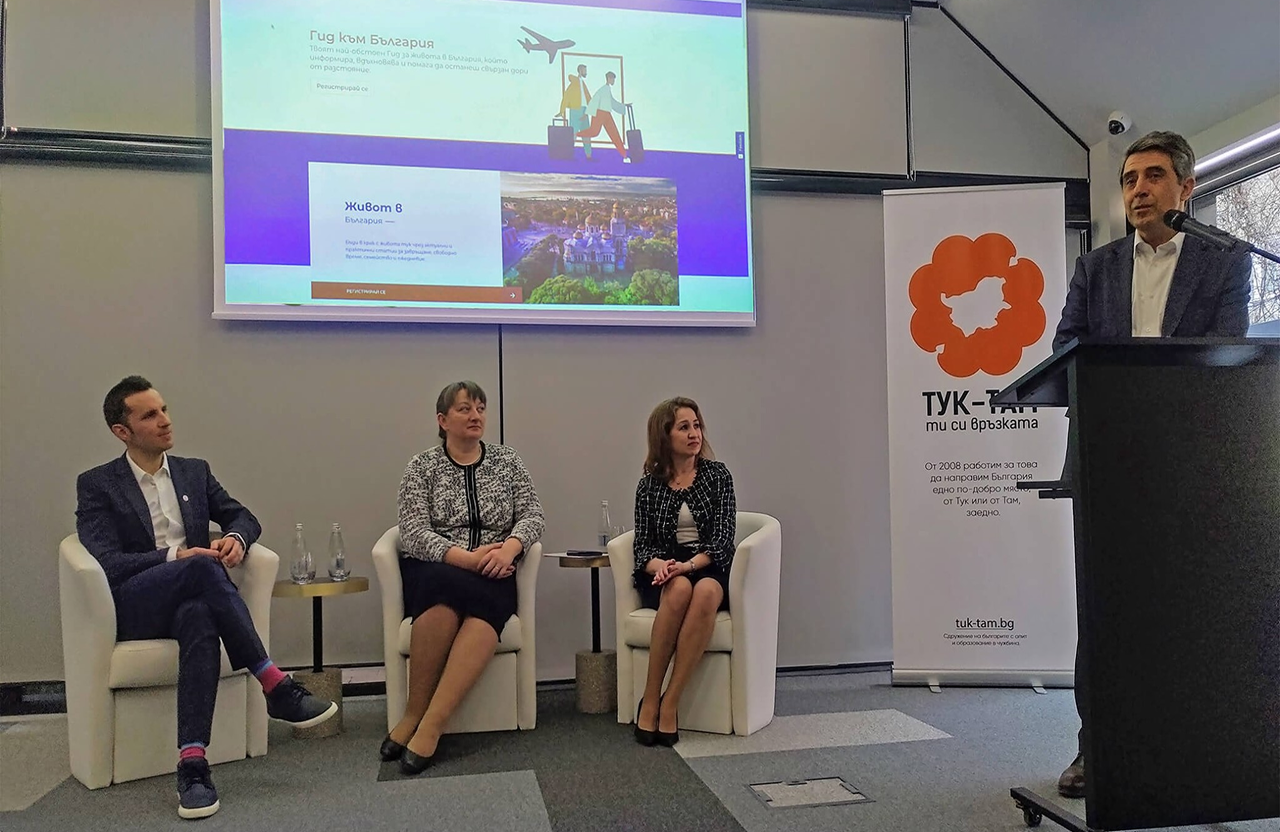 Four people at an event with three seated and one standing speaking at a podium in front of a screen.