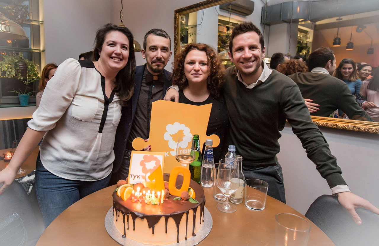 Four people smiling behind a chocolate birthday cake with orange decorations and lit candles showing number 10.