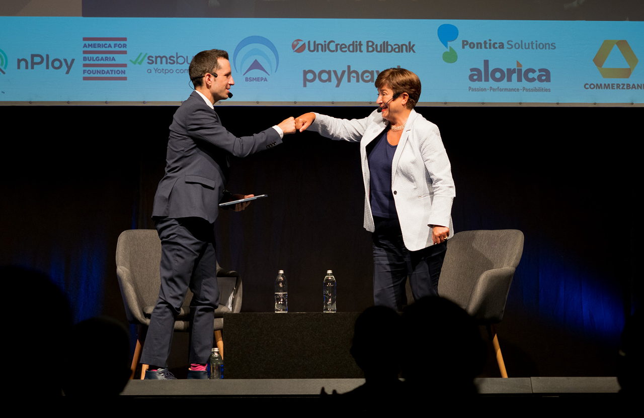 Two people on stage in suits fist bumping, with chairs and water bottles behind them.