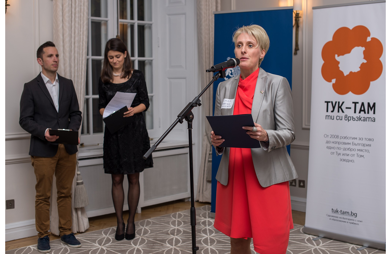 Woman in red dress and gray blazer speaking into microphone at an event with two people holding clipboards behind her.