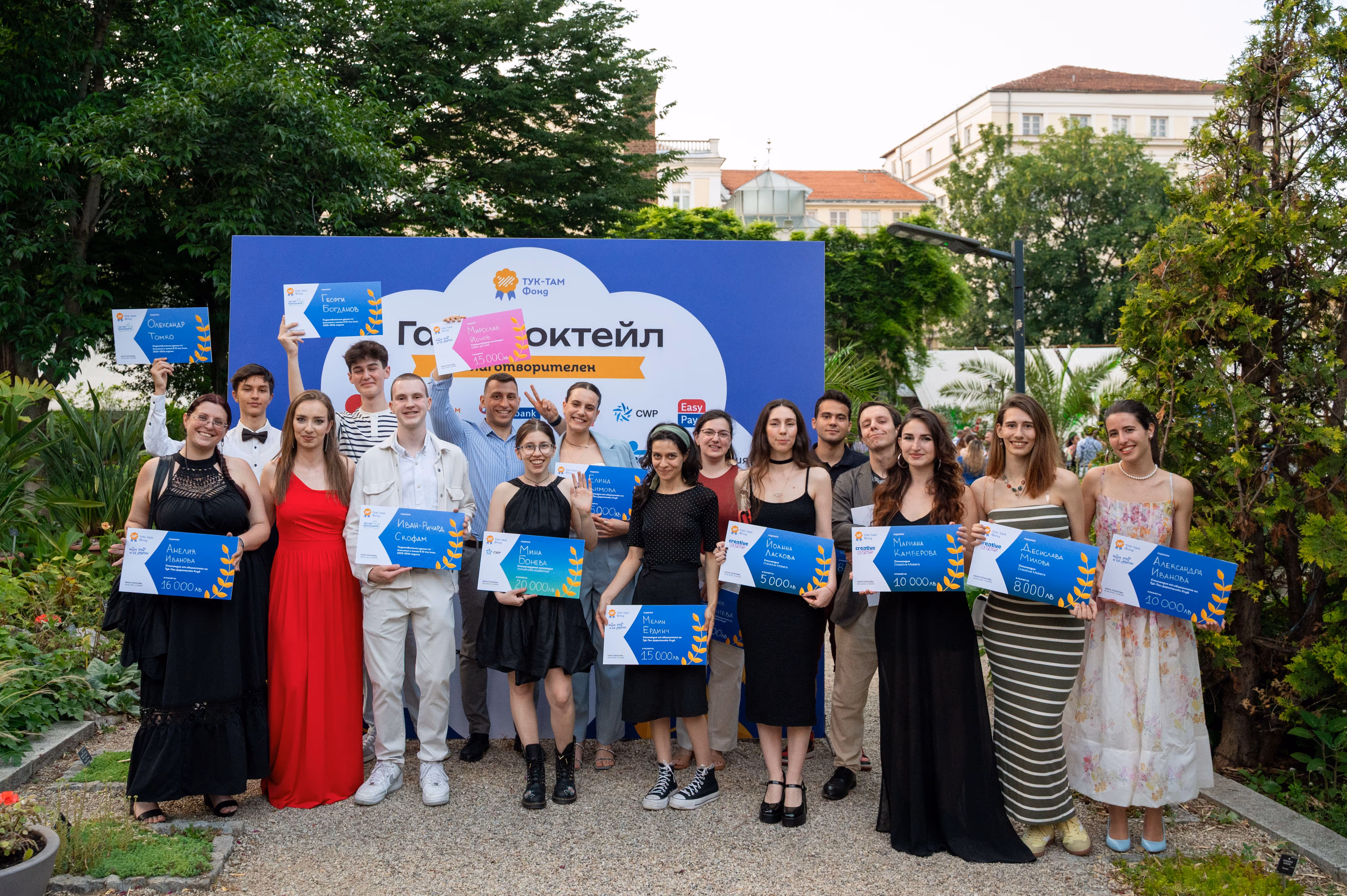 Group of young people holding scholarship checks posing at outdoor gala event with blue and white backdrop.