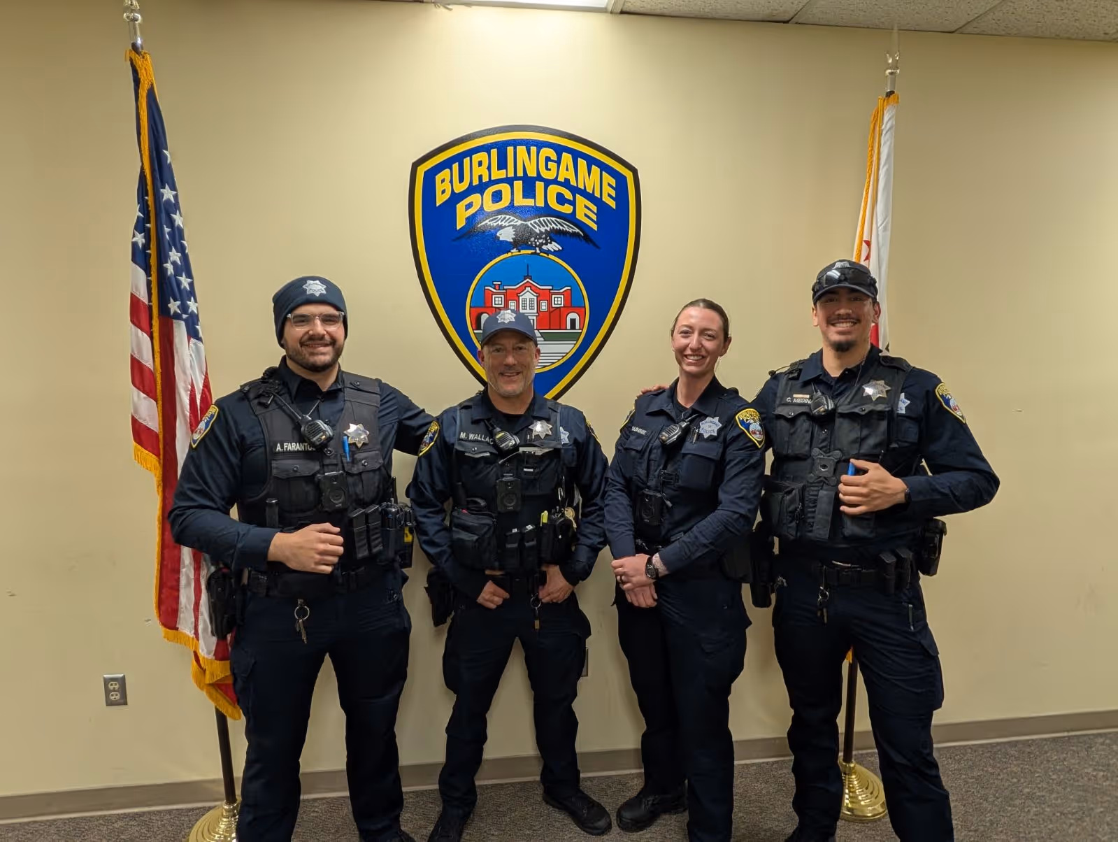 Four police officers in uniform standing and smiling in front of a Burlingame Police emblem with American and California flags on either side.