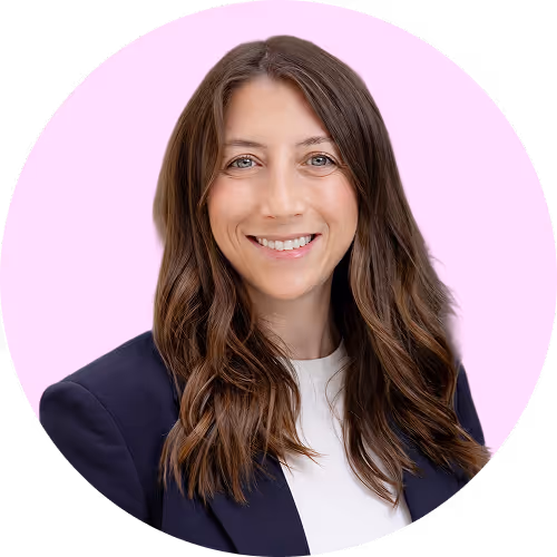 Smiling woman with long brown hair wearing a dark blazer and white top on a light pink background.