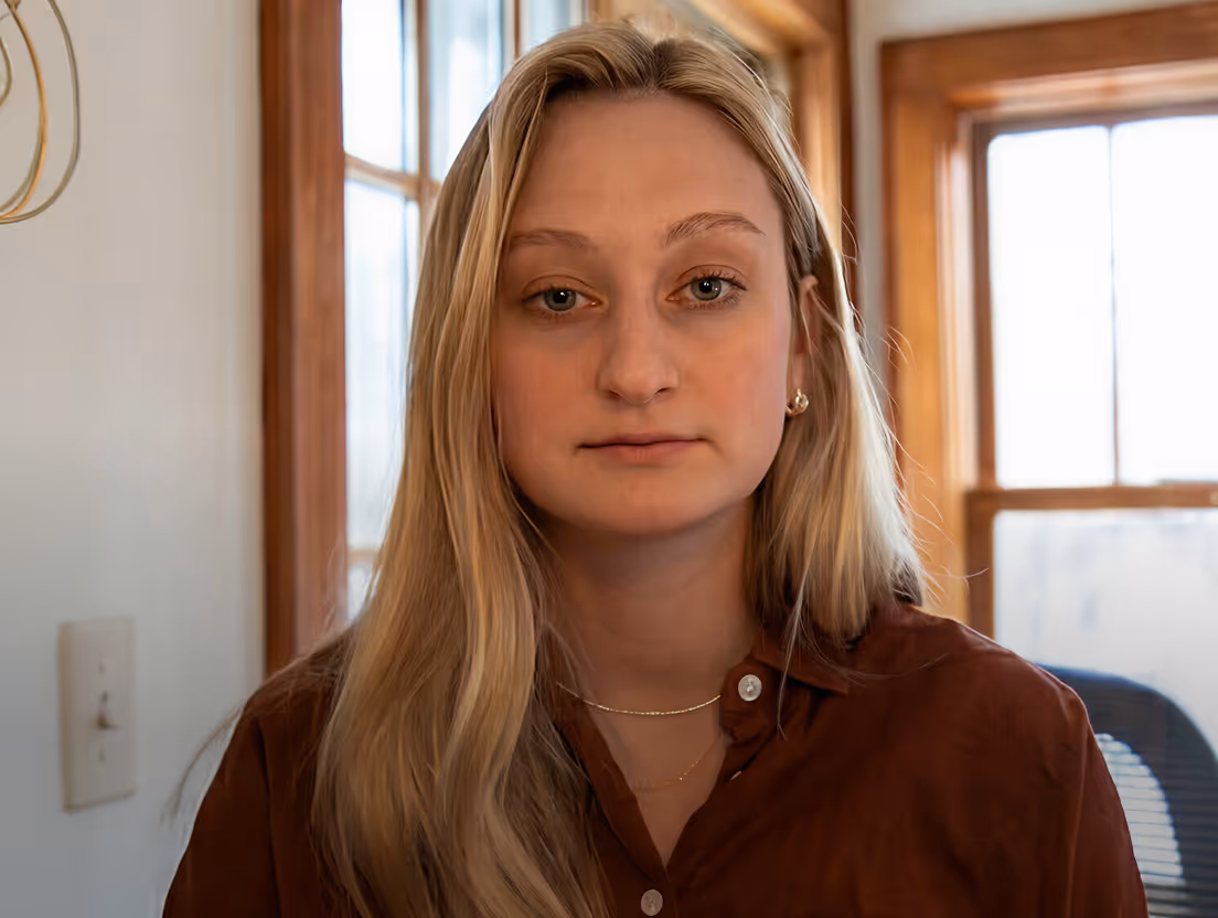 Blonde woman with long hair wearing a brown shirt and gold necklace, sitting indoors near windows.