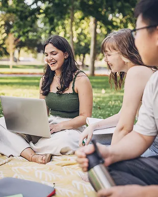 Three young adults sitting on a blanket in a park, engaging with a laptop and smiling.