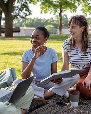 Two young women sitting on a blanket in a park, smiling and holding notebooks while a third person works on a laptop nearby.