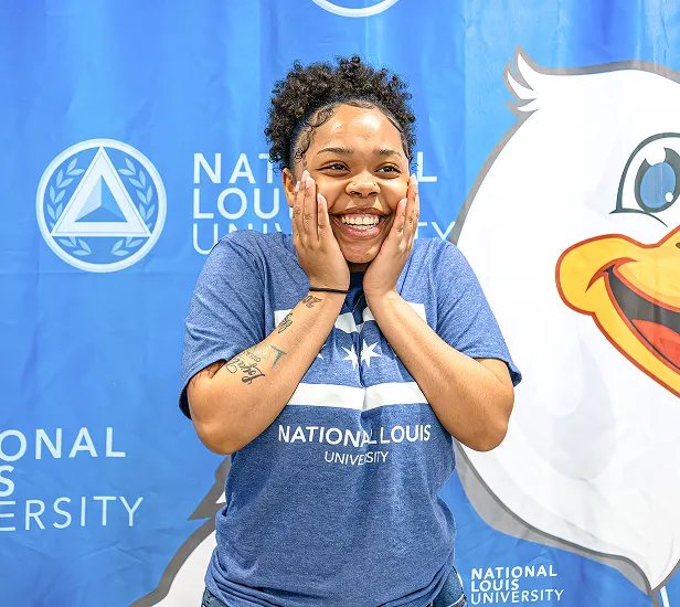 Smiling woman with curly hair, wearing a blue National Louis University shirt, standing in front of a blue backdrop with the university's logo and a large cartoon duck mascot.