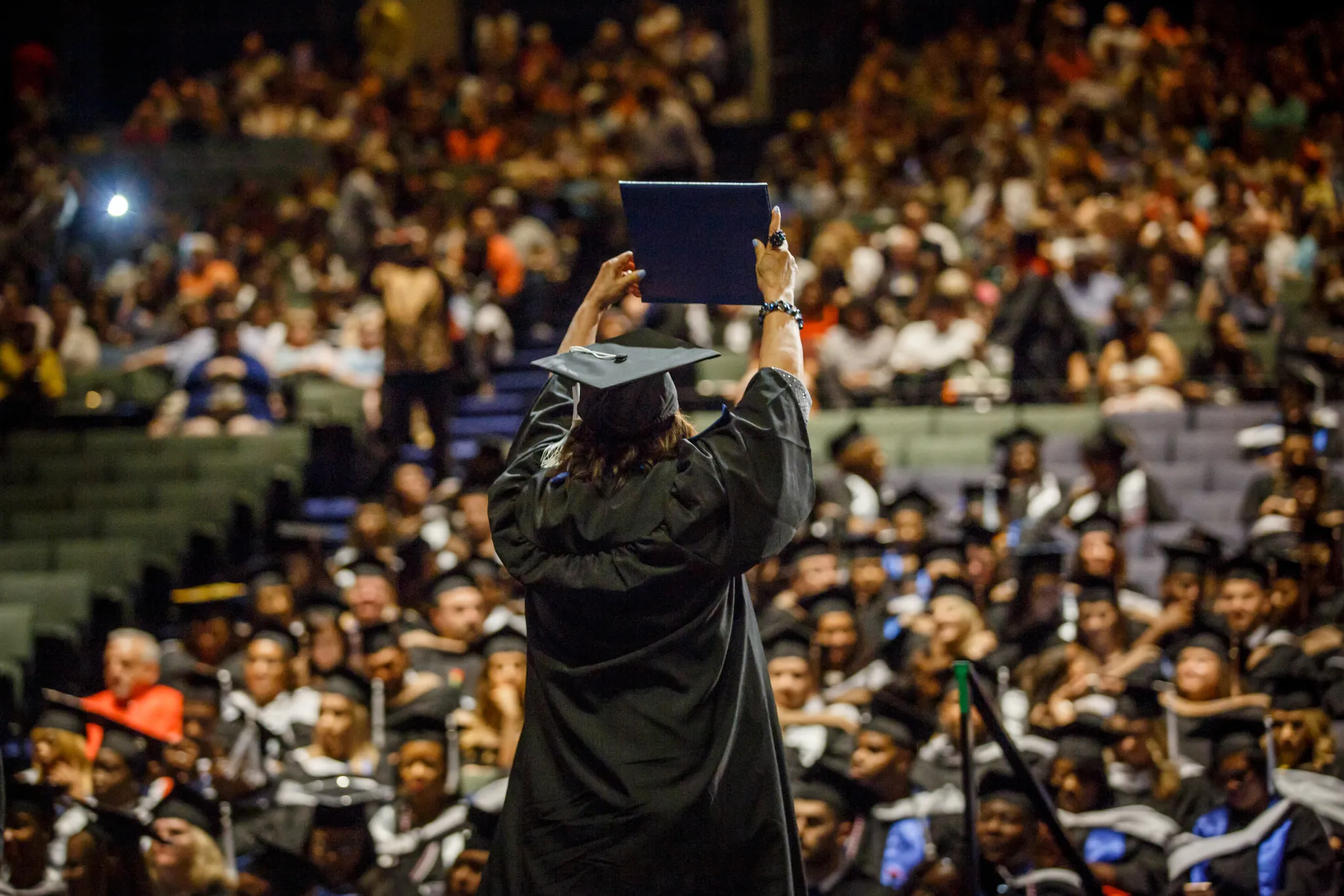 Graduate in cap and gown holding diploma up towards a crowd at a graduation ceremony.