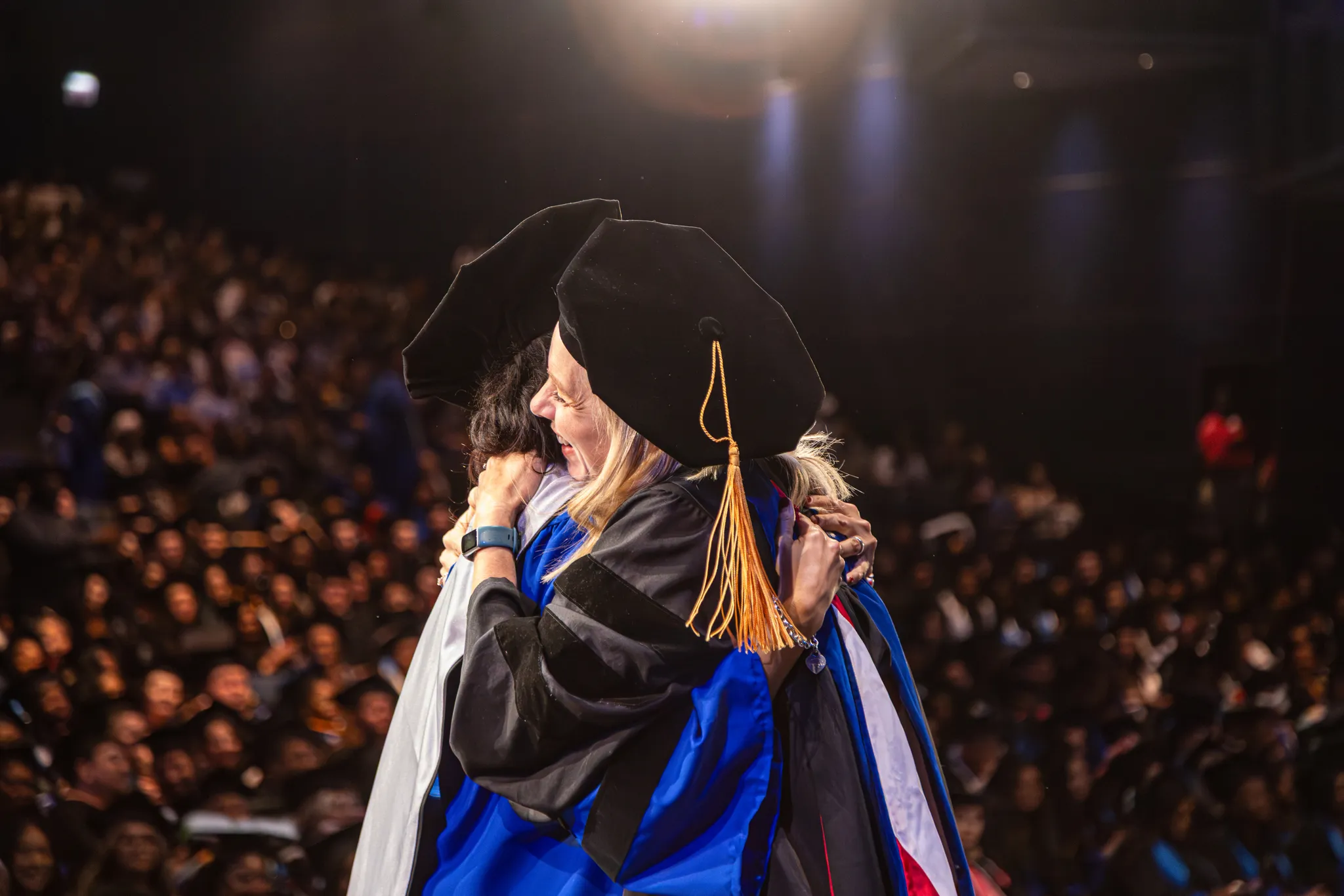 Two graduates wearing caps and gowns embracing in a crowded auditorium.