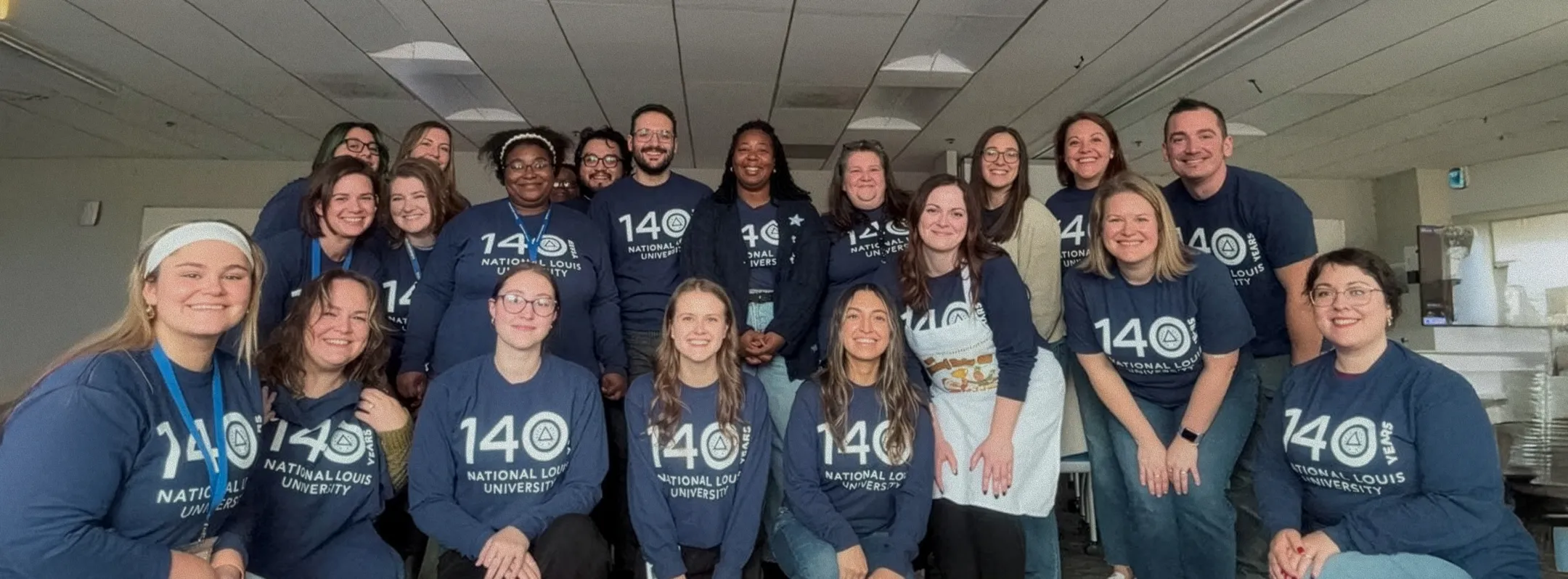 Group of people smiling indoors wearing matching navy blue shirts celebrating 140 years of National Louis University.