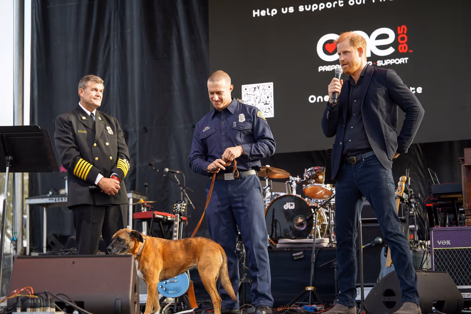 Three men on stage, one in fire chief uniform, one in firefighter uniform holding a dog on leash, and a man with red hair speaking into a microphone.
