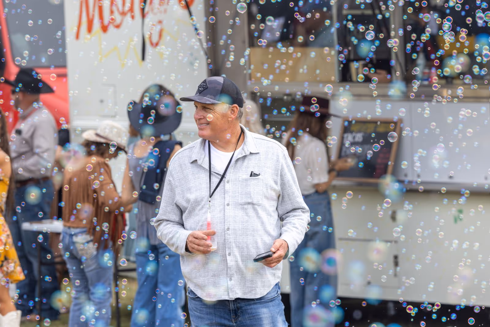 Smiling older man in a baseball cap and light shirt holding a drink and phone, surrounded by floating bubbles at an outdoor event with people in the background.