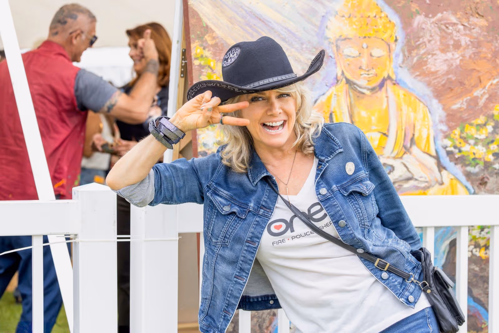Smiling woman in a black cowboy hat and denim jacket posing with a peace sign in front of a colorful Buddha painting.