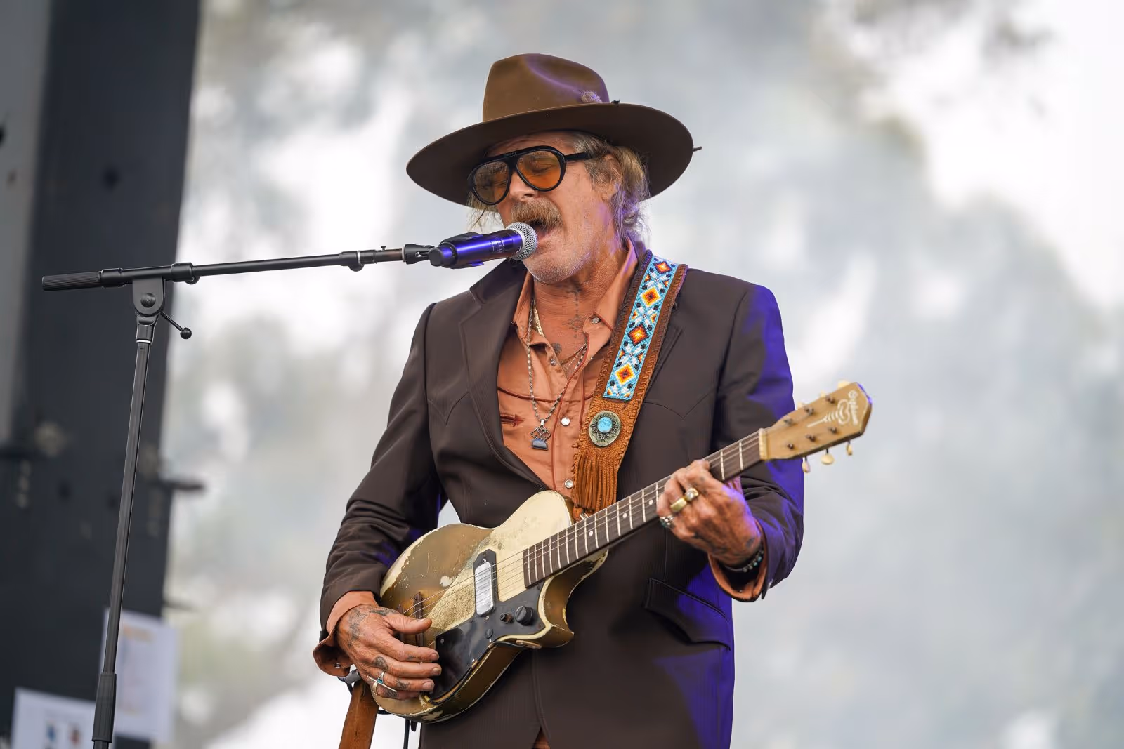 Musician wearing a brown hat and suit playing an electric guitar and singing into a microphone.
