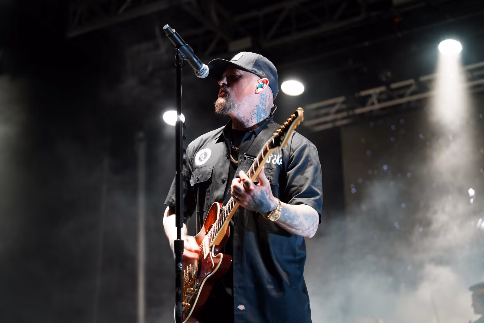 Man with tattoos wearing a cap playing electric guitar and singing into a microphone on stage with spotlights.