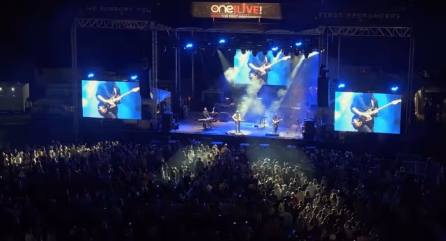 Nighttime outdoor concert with a band performing on stage and large screens showing close-ups of a guitarist, with a crowd watching.