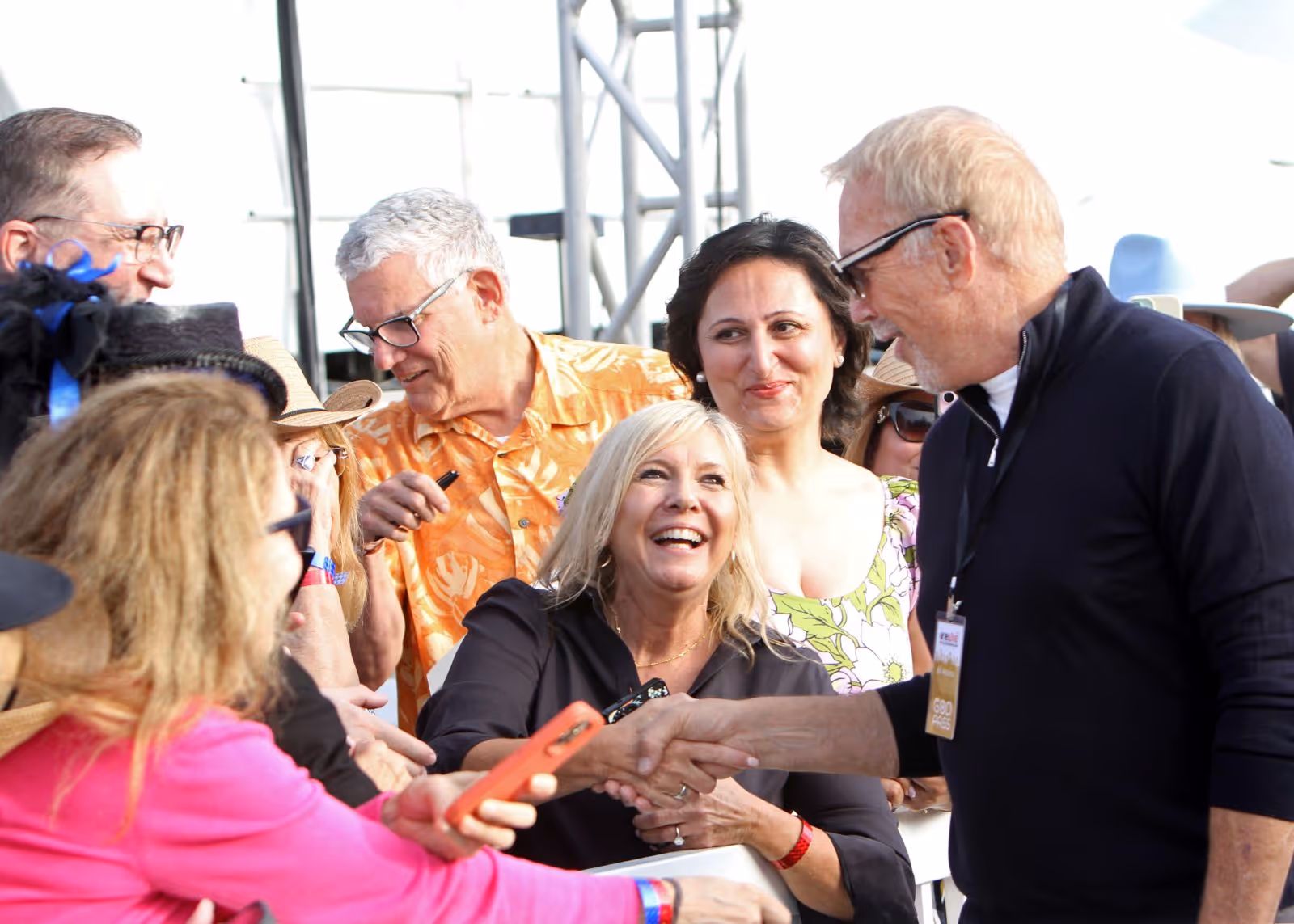 A smiling woman shakes hands with a man wearing glasses and a black jacket while other people look on and smile.