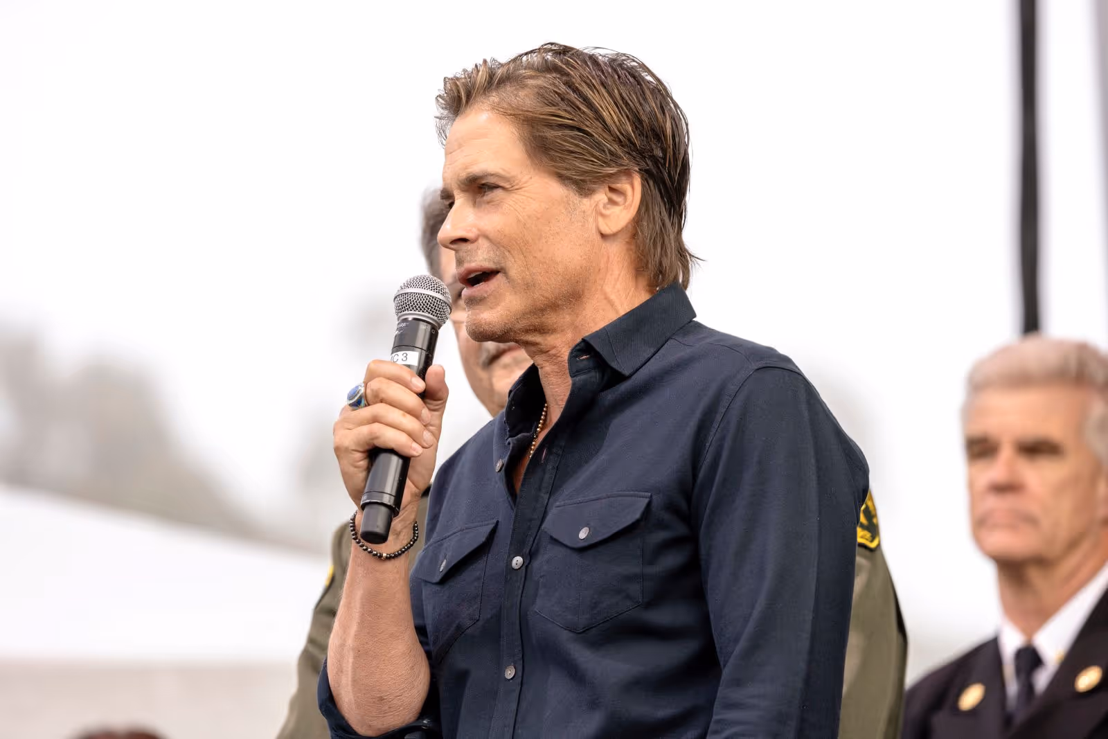 Man in a dark blue shirt speaking into a microphone at an event with uniformed individuals in the background.