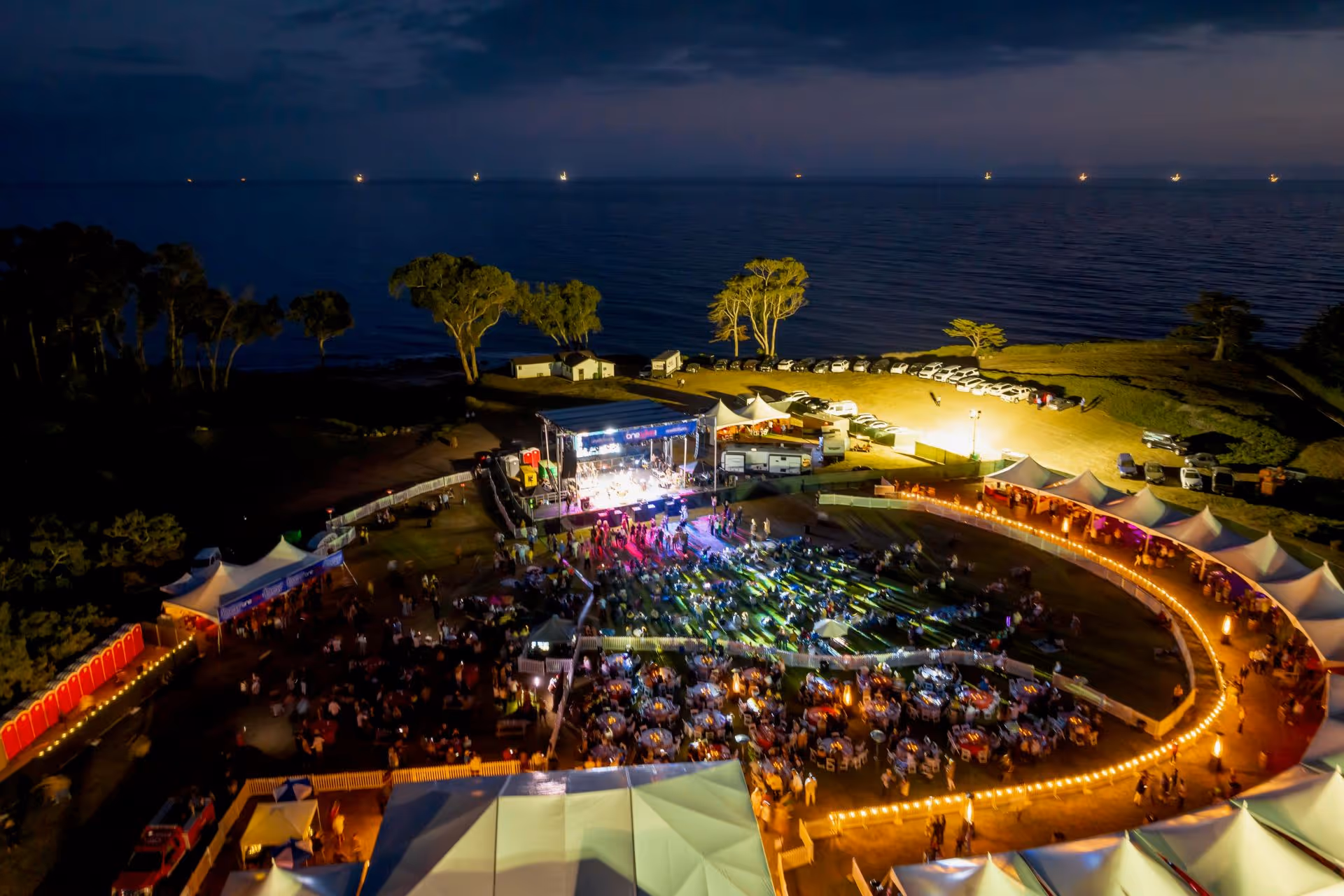 Aerial night view of a seaside outdoor event with a lit stage, seated crowd, white tents, and parked cars near the water.