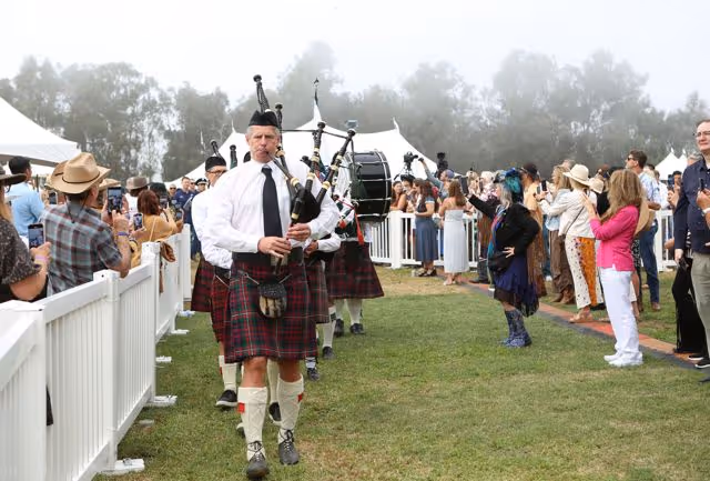 A group of bagpipers in traditional Scottish kilts playing and marching on grass while spectators watch and take photos.