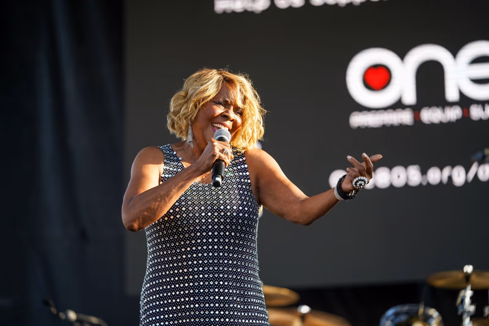 Female singer with blonde hair wearing a patterned dress, performing on stage with a microphone.