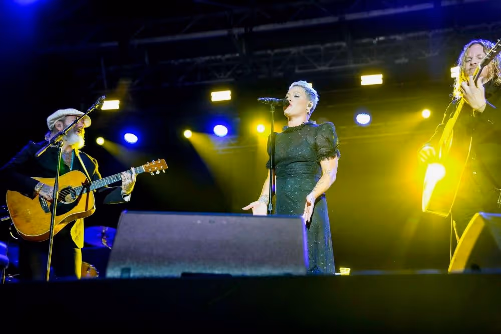 Three musicians performing on stage with two playing acoustic guitars and one singing into a microphone under bright yellow and blue lights.