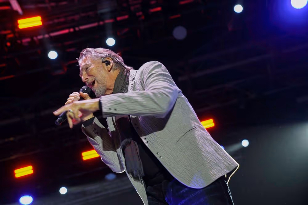 Male singer performing energetically on stage with microphone under colorful stage lights.