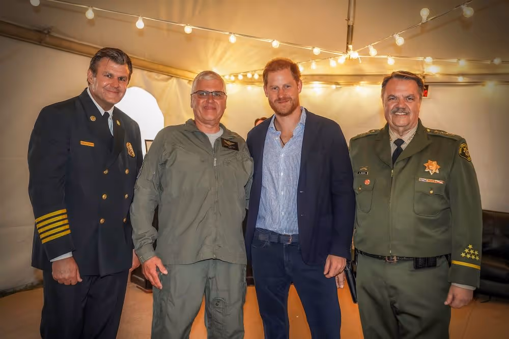 Four men standing together indoors with string lights in the background; one man is wearing a suit jacket, and the others are in various uniform styles.