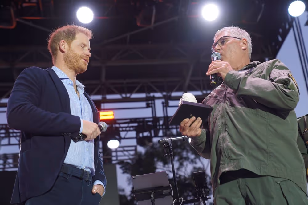 Two men on stage holding microphones, one in a suit and the other in a military-style jacket, under bright stage lights.