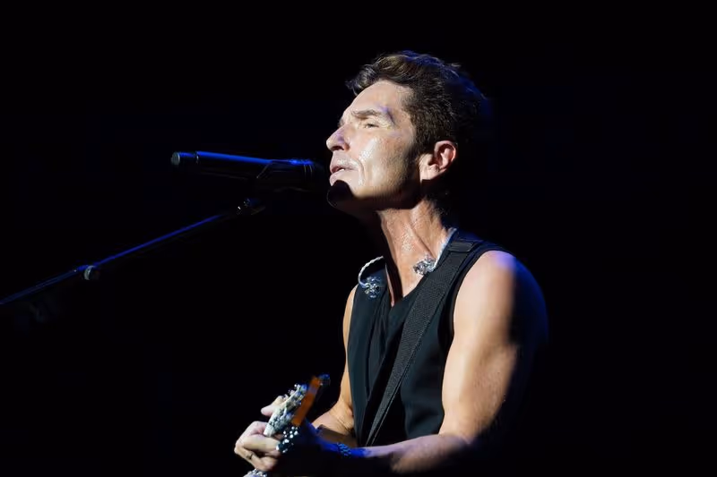 Male musician singing into a microphone and playing guitar on stage with a black background.