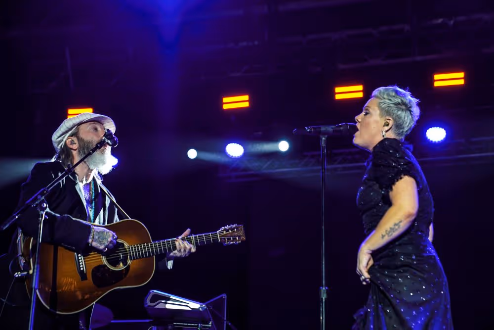 Two musicians performing on stage with a guitar and singing into microphones under blue and orange stage lights.