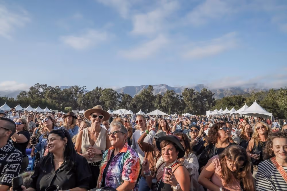 Large crowd of people enjoying an outdoor festival with white tents and mountains in the background under a partly cloudy sky.