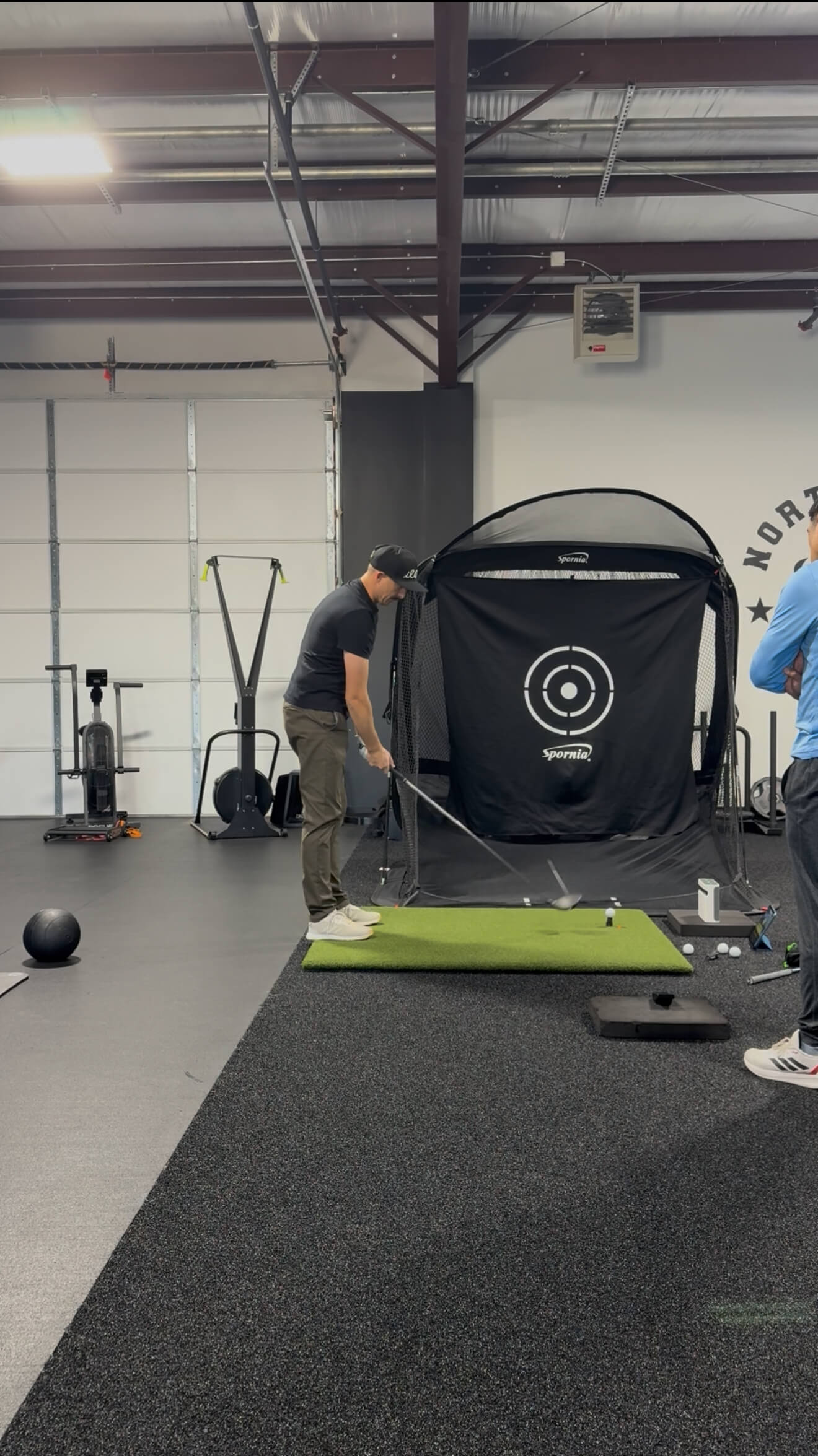 Man in a black shirt and cap practicing golf swing indoors on a green mat with a golf net target labeled Spornia in the background.