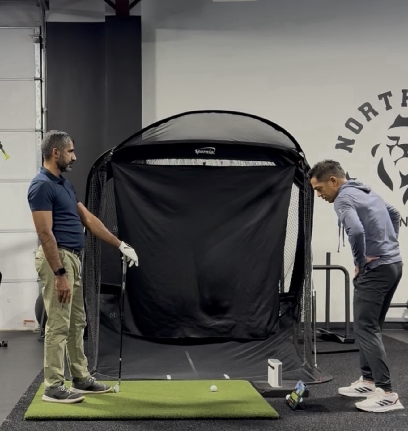 Two men in an indoor golf training area with a golf net, one holding a club preparing to hit a ball on a green mat while the other watches.