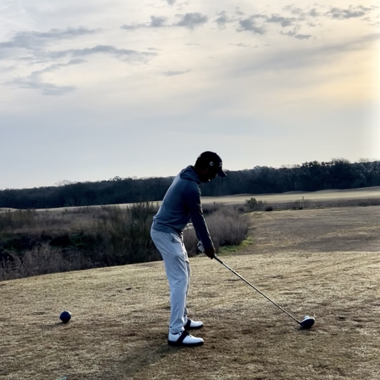Golfer in a cap and gray jacket preparing to hit a tee shot on a grassy course at sunset.