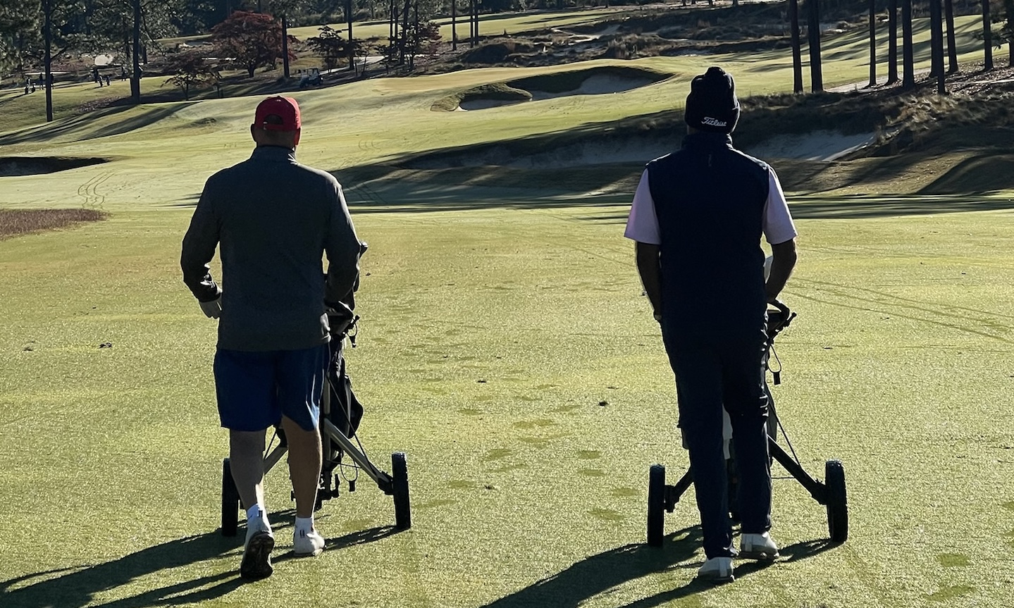 Two golfers walking on a golf course pushing golf trolleys toward a green with sand bunkers.