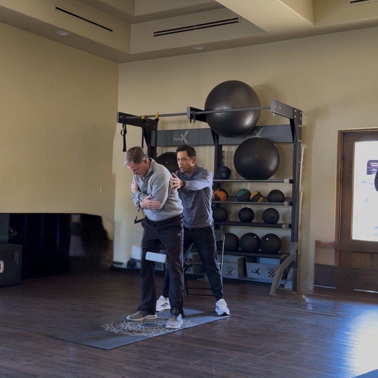 A fitness trainer assists an older man performing a stretching exercise on a yoga mat in a gym with exercise balls in the background.
