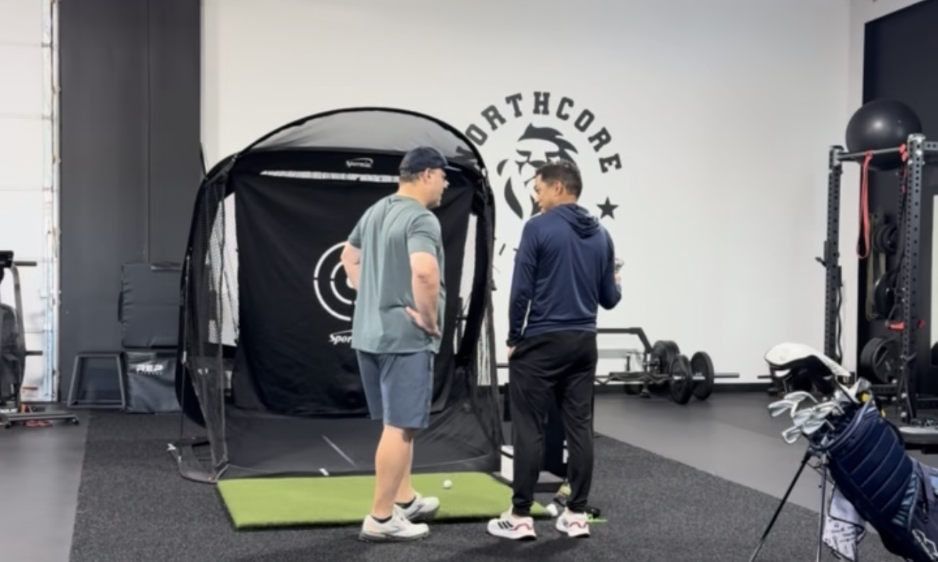 Two men standing and talking inside a sports training facility with a golf practice net, golf clubs, and exercise equipment visible.