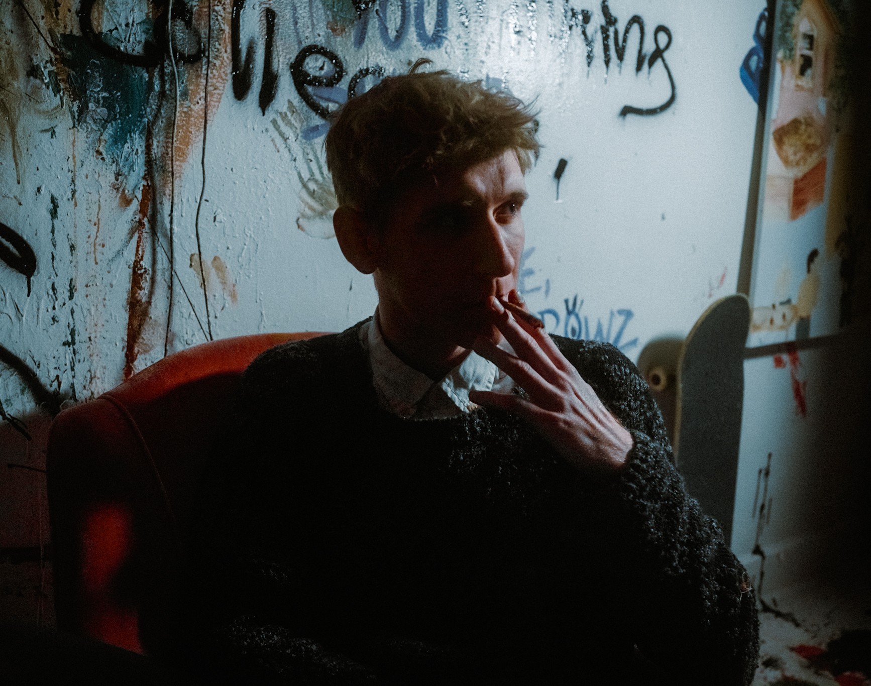 William Carroll seated on a red chair, holding a cigarette in a dimly lit room with graffiti on the walls.

Transcribed Text:

[Partial graffiti with no clear transcription available]