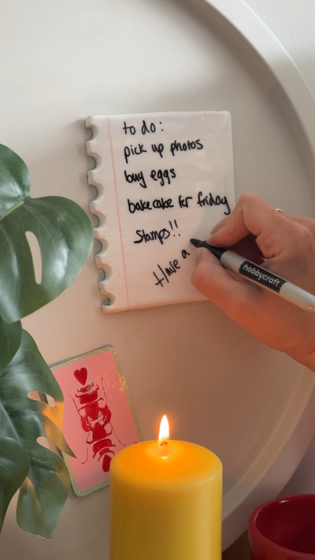 Hand writing on a handmade clay notepad-style memo board with a black marker, displaying a to-do list on a decorative tray beside a candle and plant.