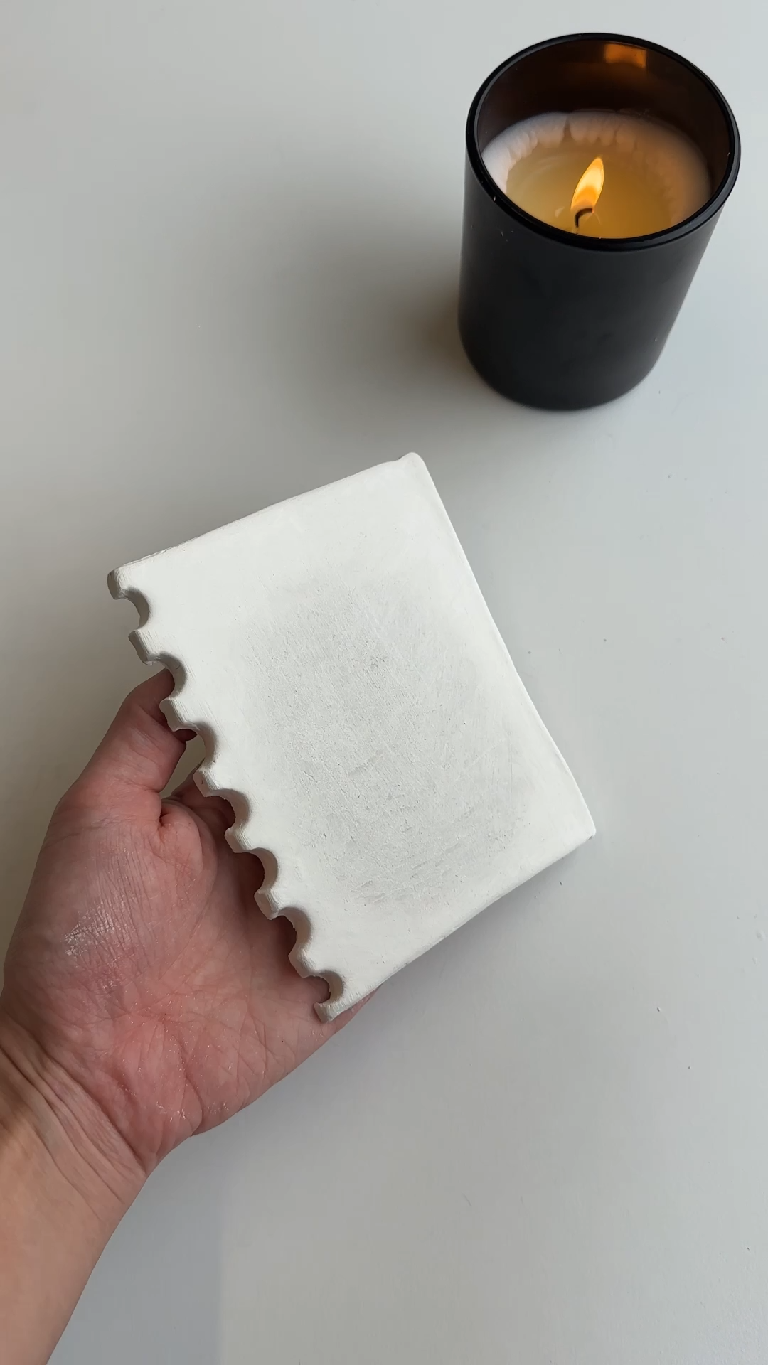 Hand holding a handmade air dry clay notepad shape with spiral-style binder indents while the clay dries on a table beside a lit candle.