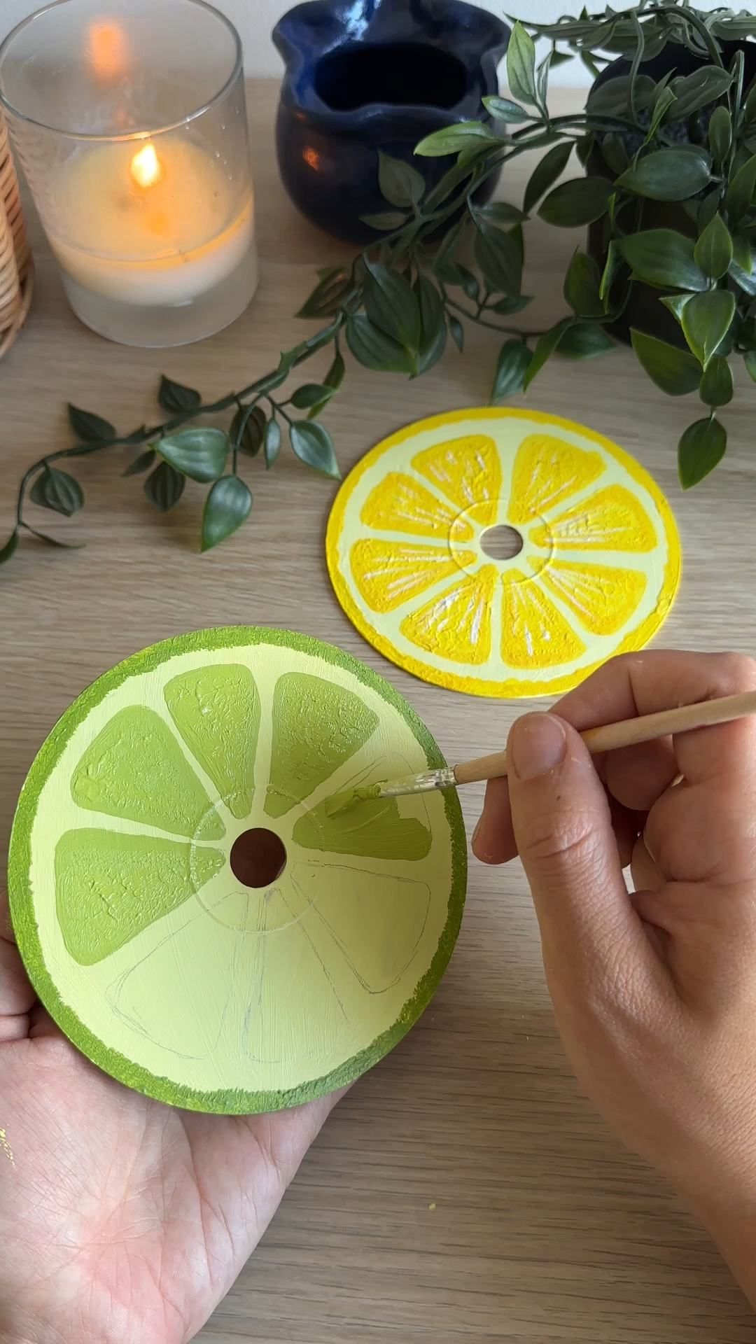 Hand painting a lime slice design onto an upcycled CD using green paint, with a finished lemon slice coaster nearby on a wooden table.