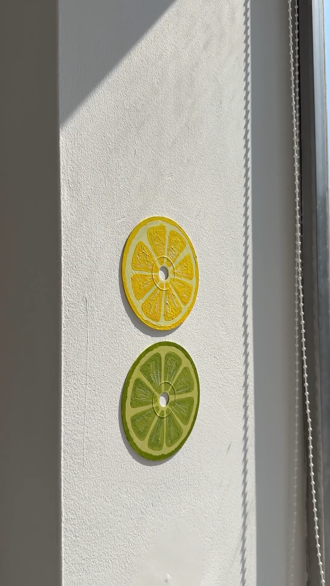 DIY citrus slice wall art made from upcycled CDs, painted as a lemon and lime and mounted on a white wall in sunlight.