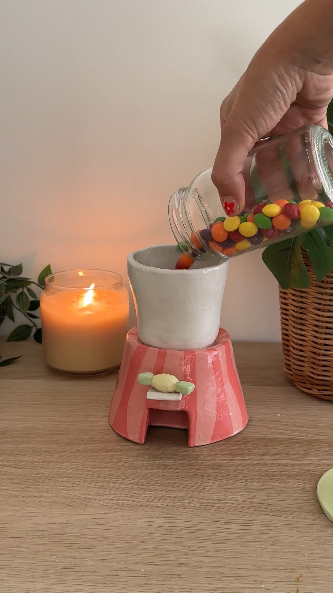 Hand pouring colourful sweets from a glass jar into a handmade ceramic sweet dispenser on a wooden table.
