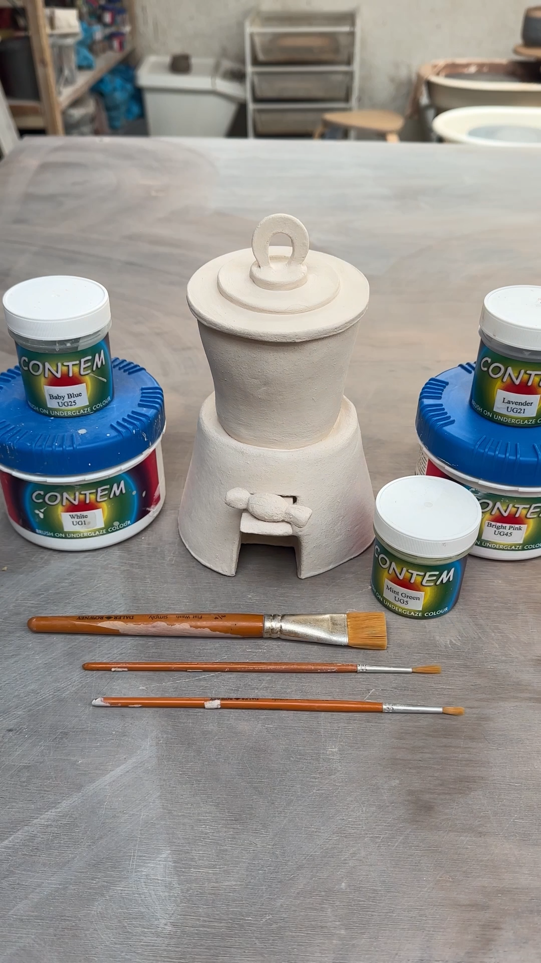 Bisque-fired ceramic sweet dispenser placed on a studio table surrounded by underglaze pots and paintbrushes, ready for decorating.