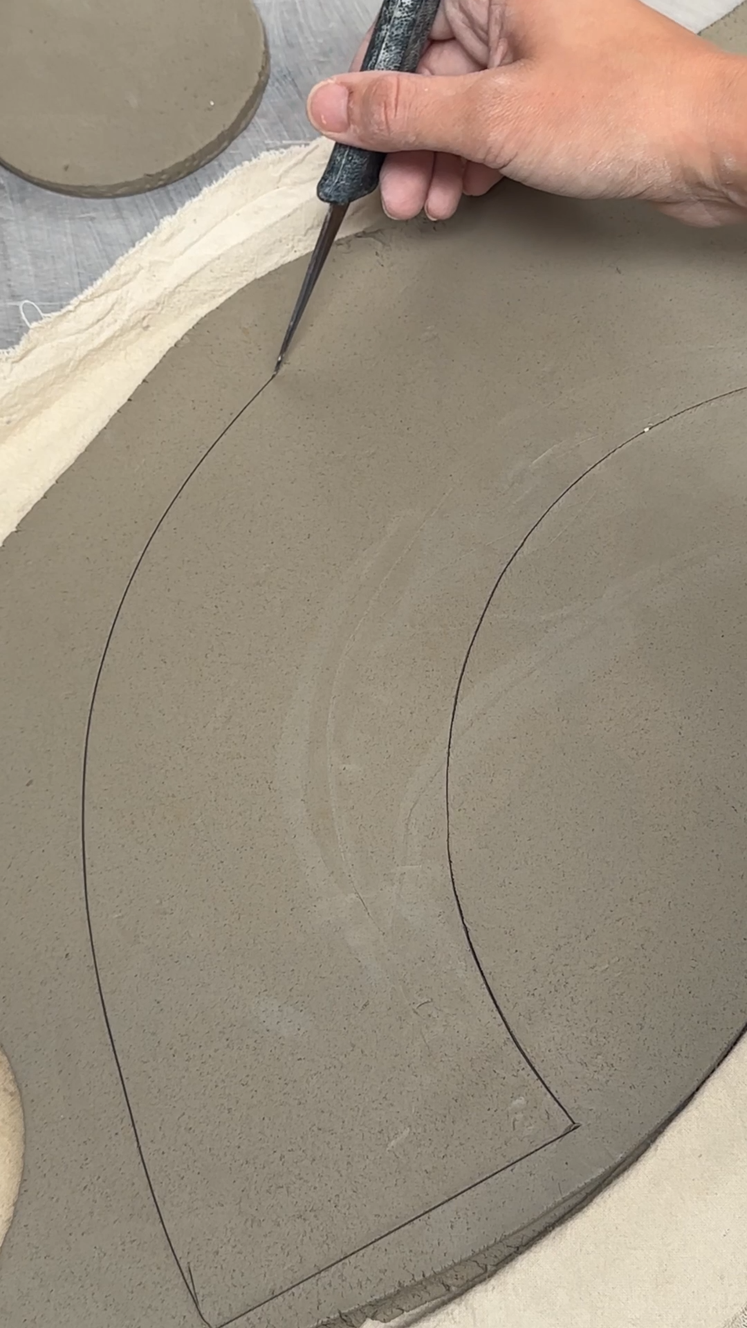 Hand using a clay needle tool to cut an arched rainbow shape from a rolled slab of clay.