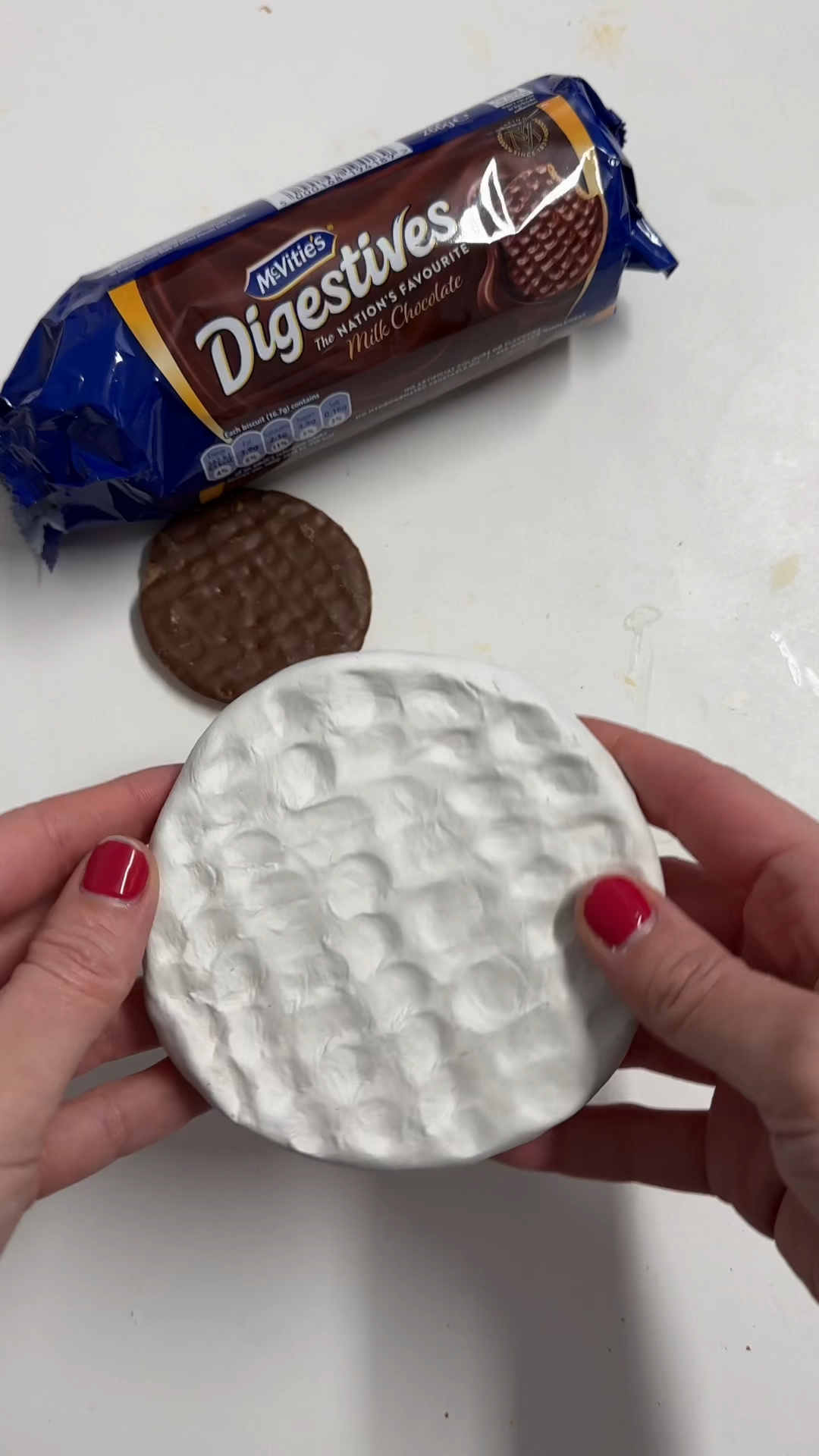 Freshly textured air dry clay biscuit coaster with ripple pattern held up before painting, next to a packet of chocolate digestives.