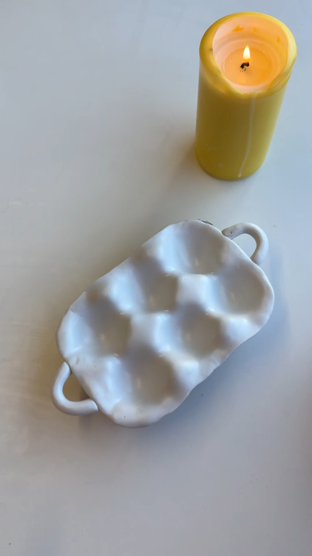 White clay egg tray with handles shaped like an egg carton, placed on a table beside a lit yellow candle.