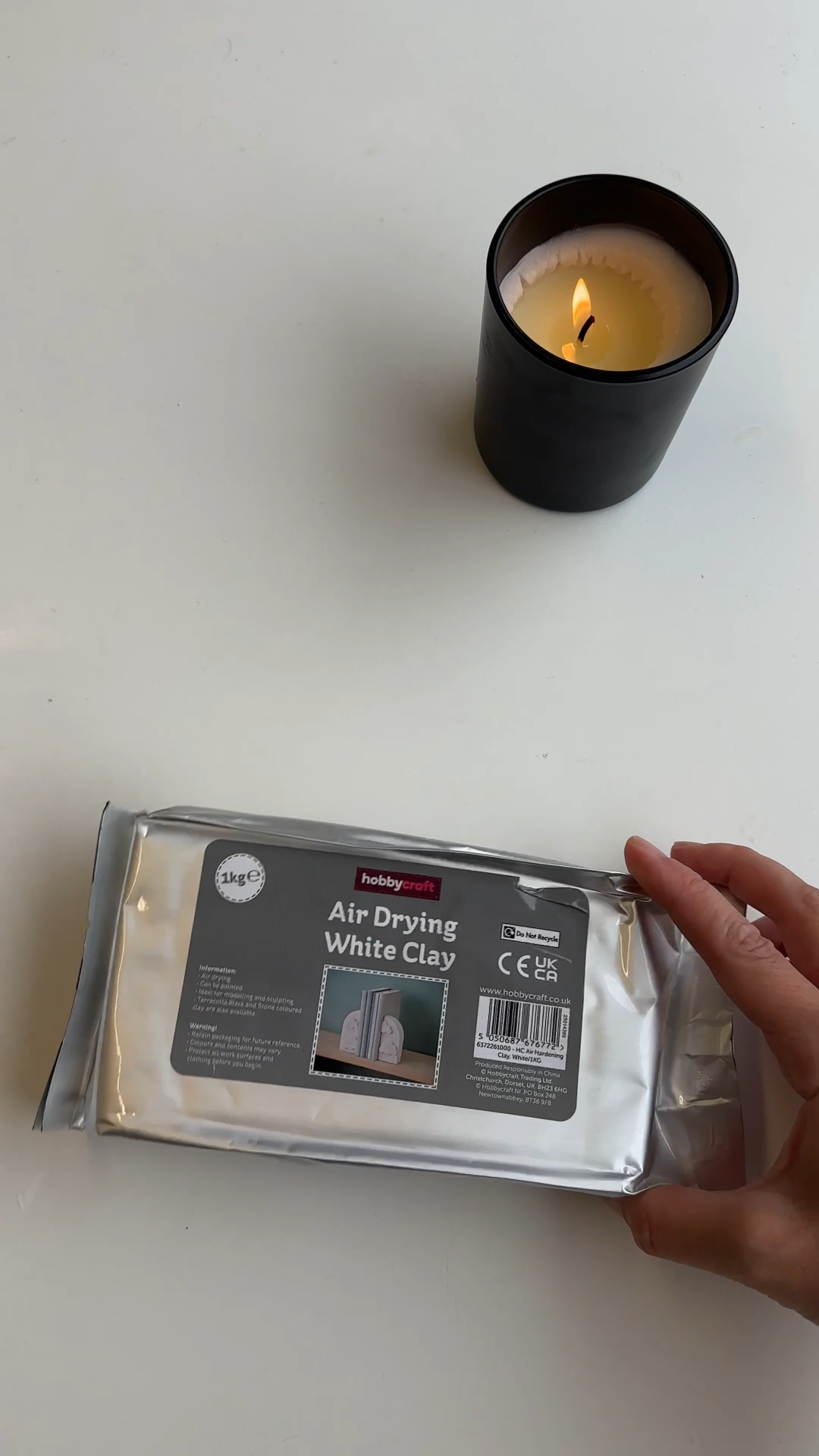 Hand holding a pack of white air-drying clay on a white table, with a lit candle in a black glass jar in the background.