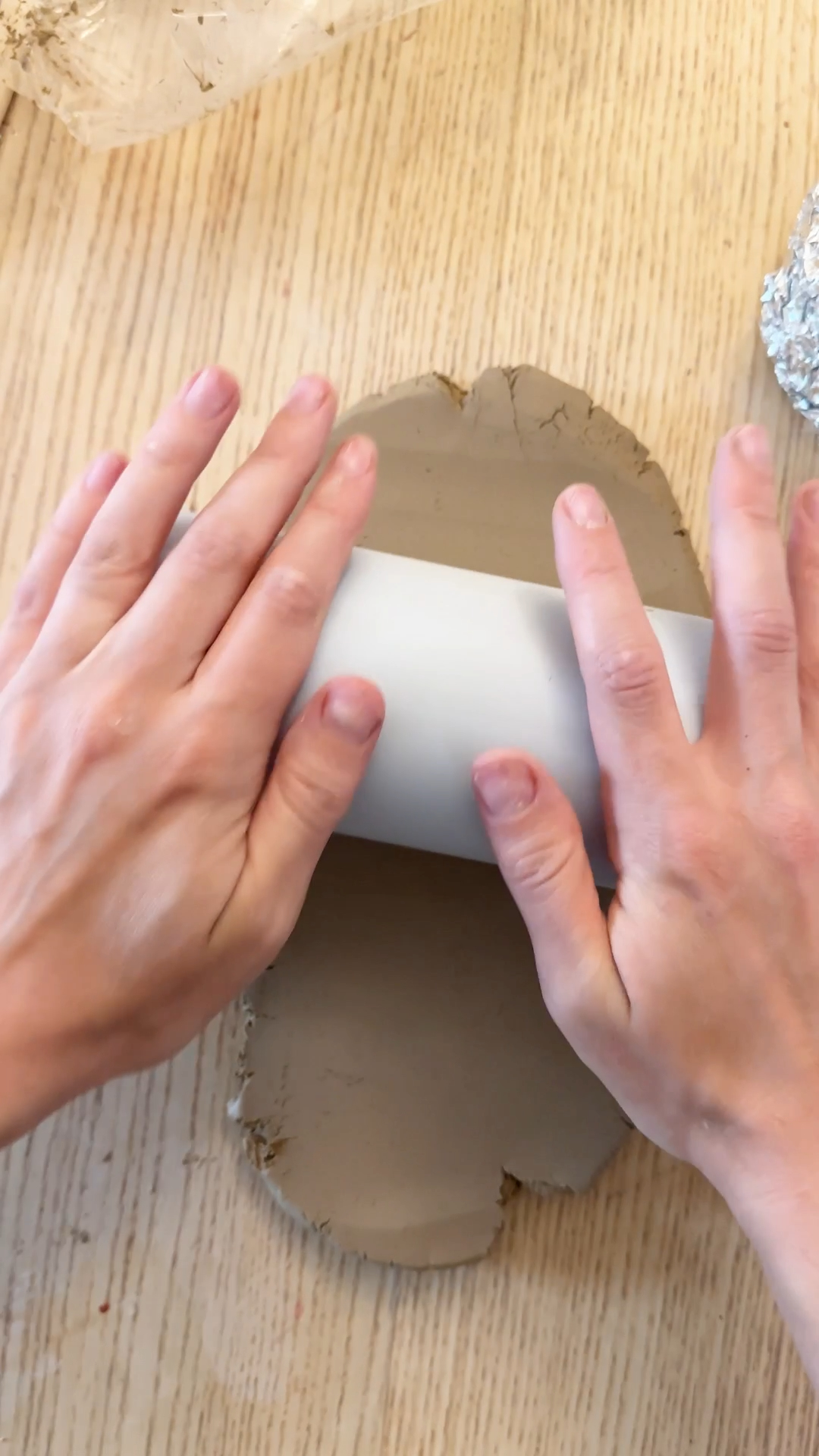Hands rolling out a slab of air dry clay on a wooden table using a water bottle as a makeshift rolling pin.